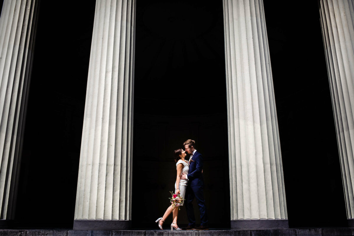 Bräutigam nimmt Braut vor dem Aachener Elisenbrunnen in den Arm, sie hält einen Brautstrauss und das Paar steht zwischen zwei Säulen.
